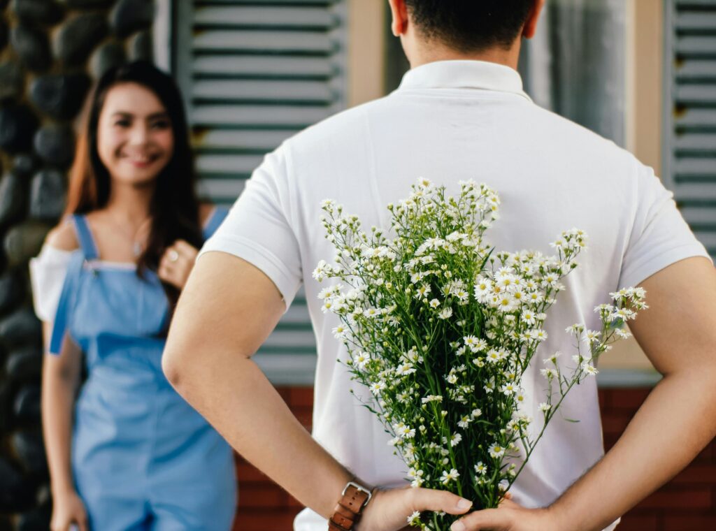 A boy faces a girl and holds flowers behind his back